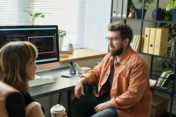 Caucasian middle aged man discussing neural network development with Caucasian young adult woman in modern office, computer monitor displaying code related to artificial intelligence