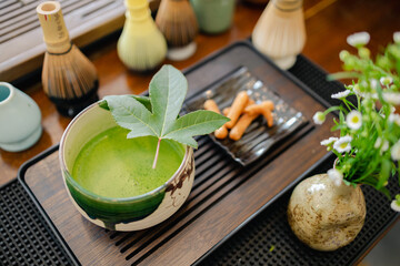 Matcha tea cup with green leaf on wooden tray with traditional bamboo whisks and small vase with flowers, peaceful tea ceremony setting with natural elements