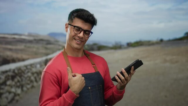 Young man wearing glasses and red sweatshirt, holds tablet smiling outdoors on a quiet city street.