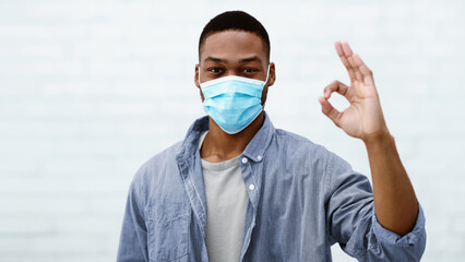 African Guy Wearing Protective Face Mask Gesturing Okay Posing Looking At Camera Standing Over Gray Wall Indoor. Covid-19 Virus, Coronavirus Protection And Prevention Healthcare Concept
