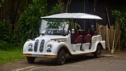 A vintage-style white and gold electric golf cart or tour vehicle with red seats is parked on a paved road surrounded by lush tropical foliage