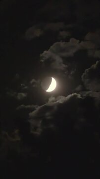 The crescent moon in the first quarter phase is hidden behind clouds in the night's black sky. Clouds move quickly in the dark sky