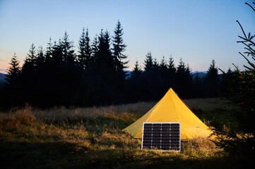 Yellow tent pitched in grassy field with solar panel beside it, capturing last light of setting sun.