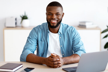 Portrait Of Young Black Freelancer Guy Sitting At Desk With Laptop Computer At Home Office, Looking At Camera, Smiling African American Entrepreneur Man Enjoying Remote Work And Self-Employment