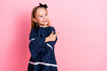 Smiling young girl in casual dress points sideways on pink background, showcasing a joyful and...