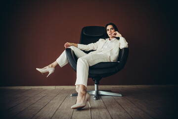 Stunning businesswoman in an elegant white suit seated stylishly in a modern chair against a brown...
