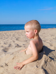 A young boy sits half-buried in sand on a sunny beach, gazing thoughtfully toward the sea. A quiet...