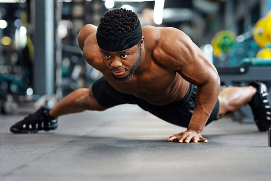 Push-up, press-up at gym concept. Motivated black guy bodybuilder doing some push-ups with one hand gym, panorama with copy space. African american shirtless muscular man working on triceps, biceps