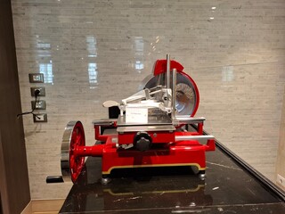 A red and silver cheese slicer sits on a countertop. The machine is large and has a black handle