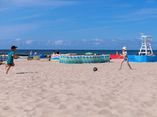 Two children play soccer on a sandy beach under a clear blue sky. One runs barefoot toward the...