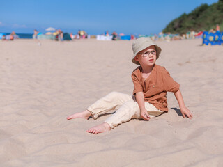 Thoughtful child sits barefoot on a sunny beach, dressed in earthy tones with a bucket hat and...