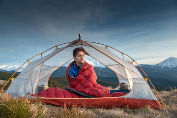 Snowy mountain landscape behind tent with male camper sitting in sleeping bag on mountain slope