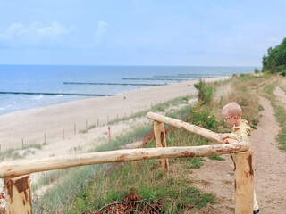 A little boy in glasses stands by a wooden fence on a coastal path, looking down thoughtfully....