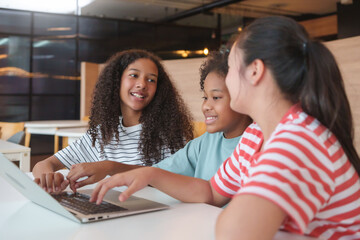 Smiling Three Diverse little girls learning together on a laptop in a classroom, showing joy,...