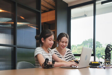 Two young Diverse girls learning to program robot arms on laptop in a stem education class, smiling...