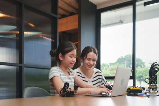 Two young Diverse girls learning to program robot arms on laptop in a stem education class, smiling and studying coding skills in a modern indoor technology