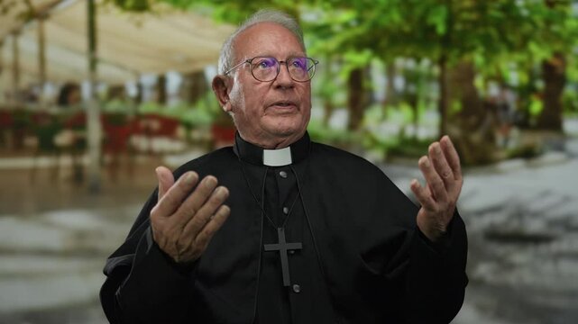 Senior priest man wearing glasses gestures outdoors on a street, with blurred green trees and people in the background, conveying a sense of peaceful spirituality.