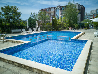 Clean outdoor pool with clear blue water, sun loungers, and umbrellas near modern apartment building. Peaceful summer vibe and relaxation area.