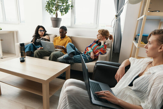 Young adult woman working on laptop in casual workplace with colleagues and friends socializing, representing Gen Z business and modern work environment.