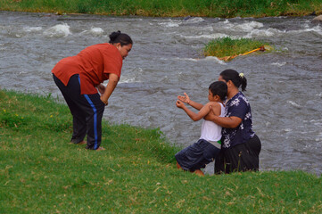 A mother lifts her young boy up from the river to the grassy riverbank, while another woman looks...
