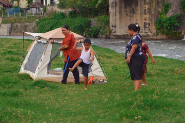 A young boy begins to show off tricks on the grassy riverside near a camping tent, while two women...