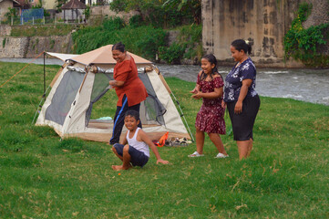 A young girl excitedly cheers on a boy performing stunts in the grass near a tent, with two women...