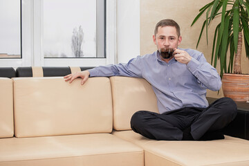 Man enjoying coffee on a beige couch while sitting cross-legged indoors on a cloudy day