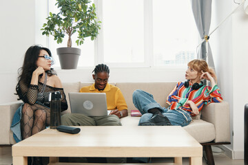young adult coworkers socializing on couch in casual workplace with laptops and headphones,...