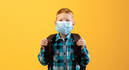 Schoolboy with backpack wearing protective face mask over yellow studio background. Redhead kid schooler using protection against coronavirus while going to school during COVID-19 pandemic