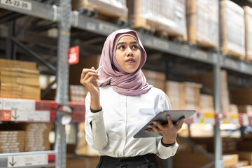 Young muslim women wearing hijab smiles while holding tablet checking goods stock inspacious warehouse. Female islamic worker at large storage facility.