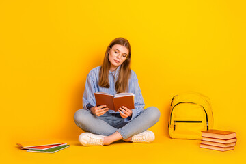 Young female student with book studying sitting cross-legged on a bright yellow background...
