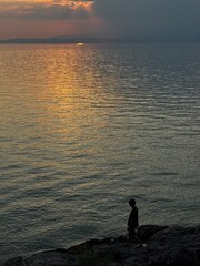 Silhouetted figure standing by the rocky coast during golden hour in Thassos, watching the sunset...