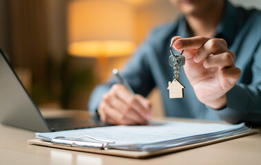 Person holds a house-shaped keychain while signing property agreement next to a miniature house...