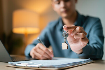 Person holds a house-shaped keychain while signing property agreement next to a miniature house...