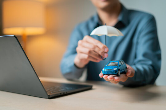 Hand holding a small blue toy car under a white umbrella beside a laptop, symbolizing online auto insurance protection.