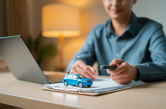 Close-up of a person holding a car key fob in one hand and a pen in the other while reviewing documents and a toy SUV on a clipboard next to a laptop, symbolizing auto insurance signing.