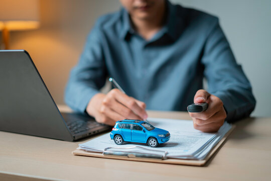 Close-up of a person holding a car key fob in one hand and a pen in the other while reviewing documents and a toy SUV on a clipboard next to a laptop, symbolizing auto insurance signing.