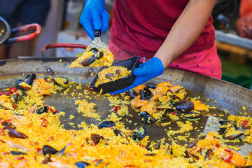 Chef in blue gloves serving seafood paella into plastic container from giant skillet during food event. Concept of paella, street food service. Spanish Culture and Food Festival