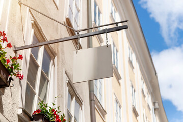 Hanging square shop sign with floral decor and sunny sky, ideal for boutique branding mockups,...