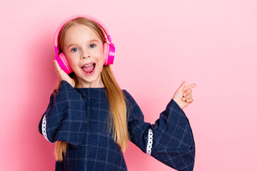 Smiling young girl wearing pink headphones and trendy dress pointing with finger against pastel...