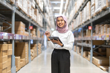 Young muslim women wearing hijab smiles while holding tablet checking goods stock inspacious warehouse. Female islamic worker at large storage facility.