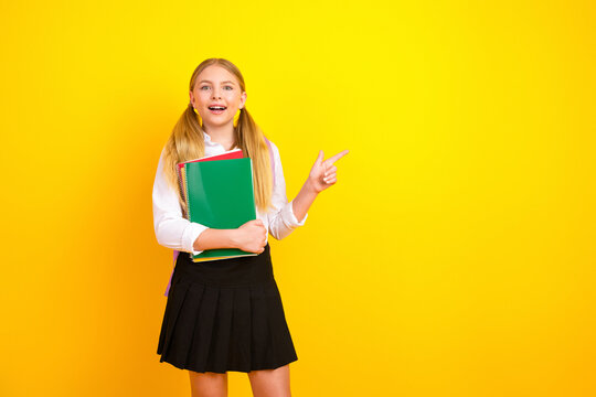 Cheerful schoolgirl holding notebooks and pointing on a vibrant yellow background, embodying educational enthusiasm