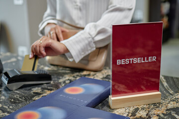 Woman paying with credit card at payment terminal near red bestseller sign on counter, holding...