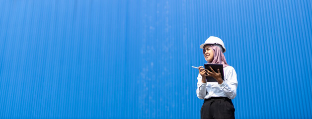 Muslim woman industrial engineer in uniform wearing safety hard hat using tablet checking containers loading. Area logistics import export and shipping cargo freight ship. Blue background.