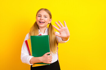 Cheerful schoolgirl holding colorful notebooks and waving with a smile against a vibrant yellow...