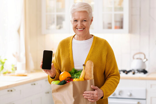 Cheerful senior woman with shopping bag full of grocery showing smartphone with blank screen, kitchen interior, copy space, mockup. Smiling old lady using mobile app for grocery delivery