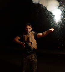 Man dressed in military clothing, posing at night with a machine gun and a lit flare illuminating...