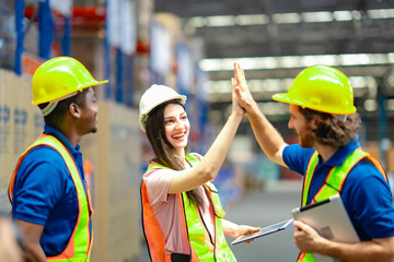 Happy logistics team giving a high five in warehouse. Workers in hard hats and safety vests display...