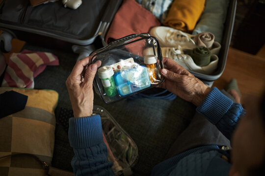 Hands of senior carefully packing medications and toiletries into clear bags for upcoming travel trip, surrounded by neatly organized suitcase and garments
