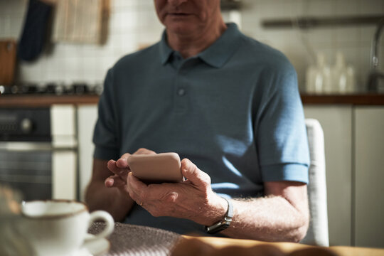 Senior man sitting in kitchen holding smartphone and concentrating on screen, with coffee cup on table in front of him